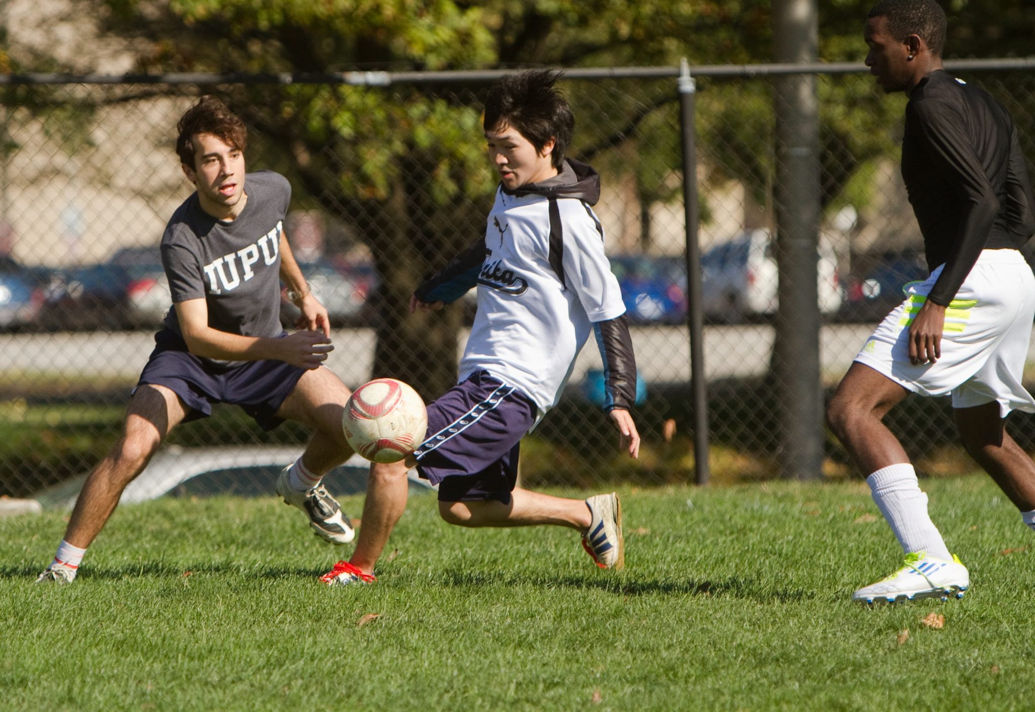 Three students playing soccer on a grassy field, with one wearing an IUPUI shirt and preparing to intercept the ball