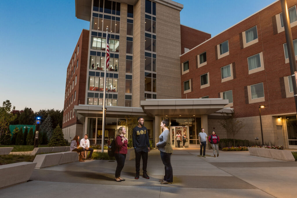 Picture of three people standing in front of a tall brick building with lots of windows as the sun is setting and there are other students around the outside of the building in the background.
